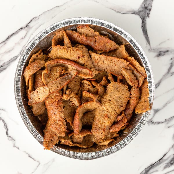 Top-down view of a foil container filled with thinly sliced, seasoned gyro meat on a marble surface.