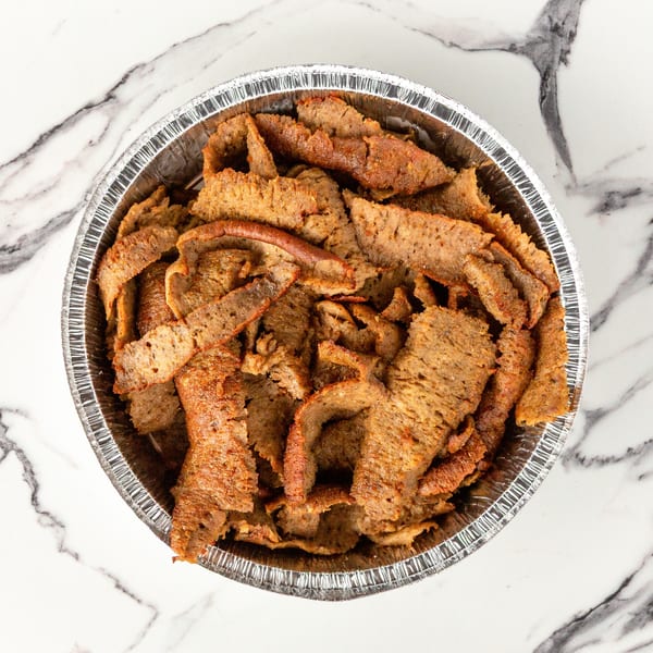 A foil container filled with thinly sliced, seasoned gyro meat on a white marble surface.