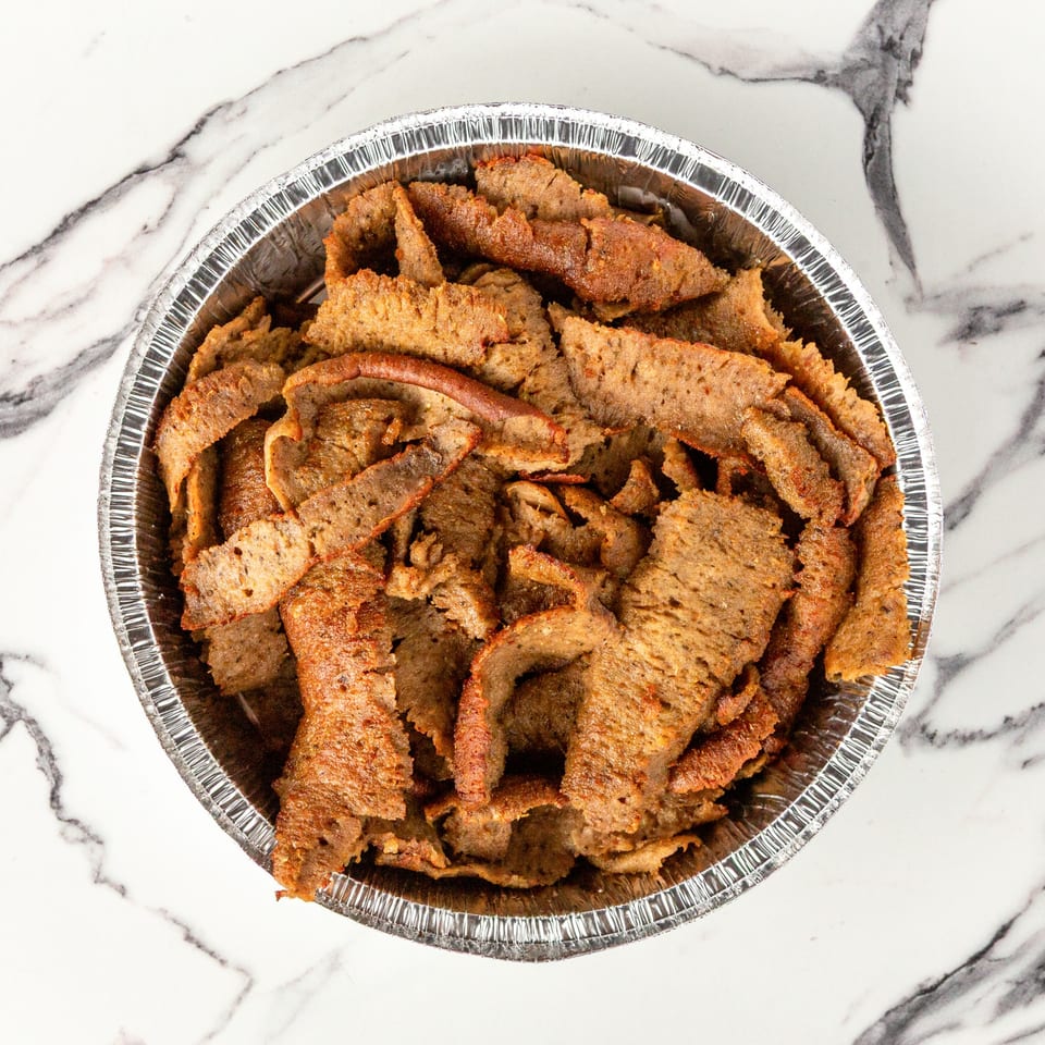 A foil container filled with thinly sliced, seasoned gyro meat on a white marble surface.