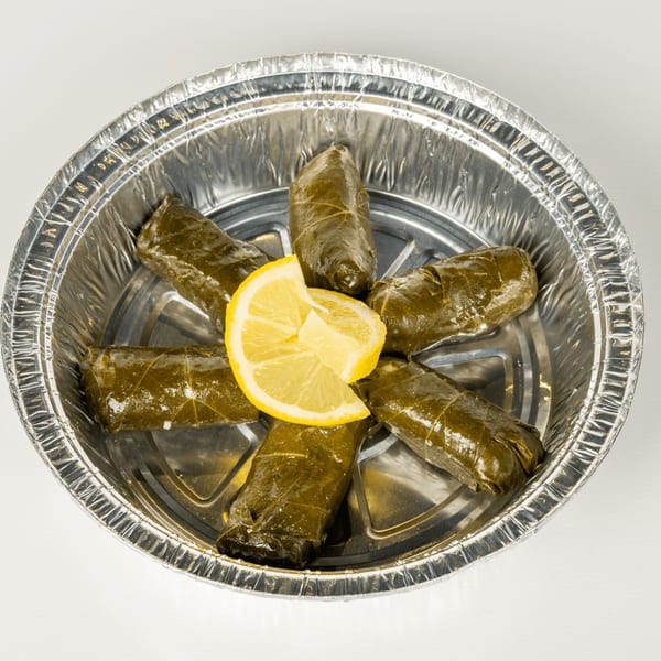 Six Greek dolmades, grape leaves stuffed with rice, arranged in a foil container with a lemon wedge.