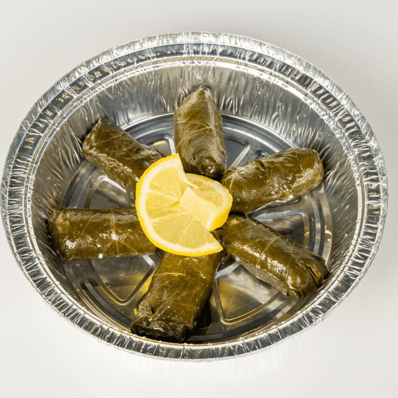 Six Greek dolmades, grape leaves stuffed with rice, arranged in a foil container with a lemon wedge.