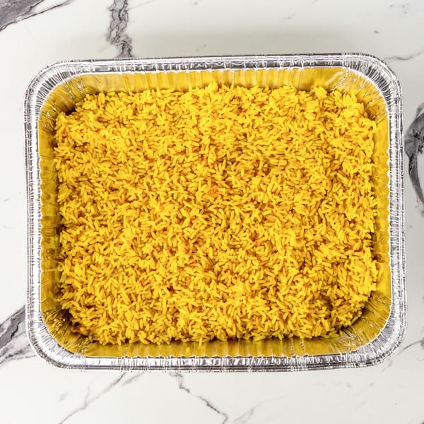 Bright yellow rice fills a rectangular aluminum foil pan, centered on a white marble surface.