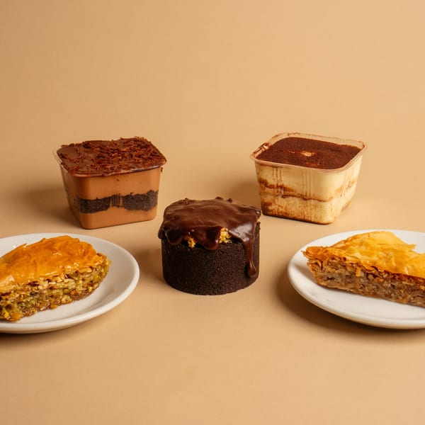 Two baklava slices on white plates, a chocolate cake, and two dessert cups on a tan background.