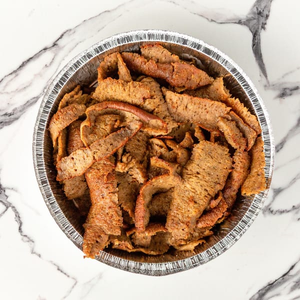 A foil container filled with thinly sliced, seasoned gyro meat on a white marble surface.