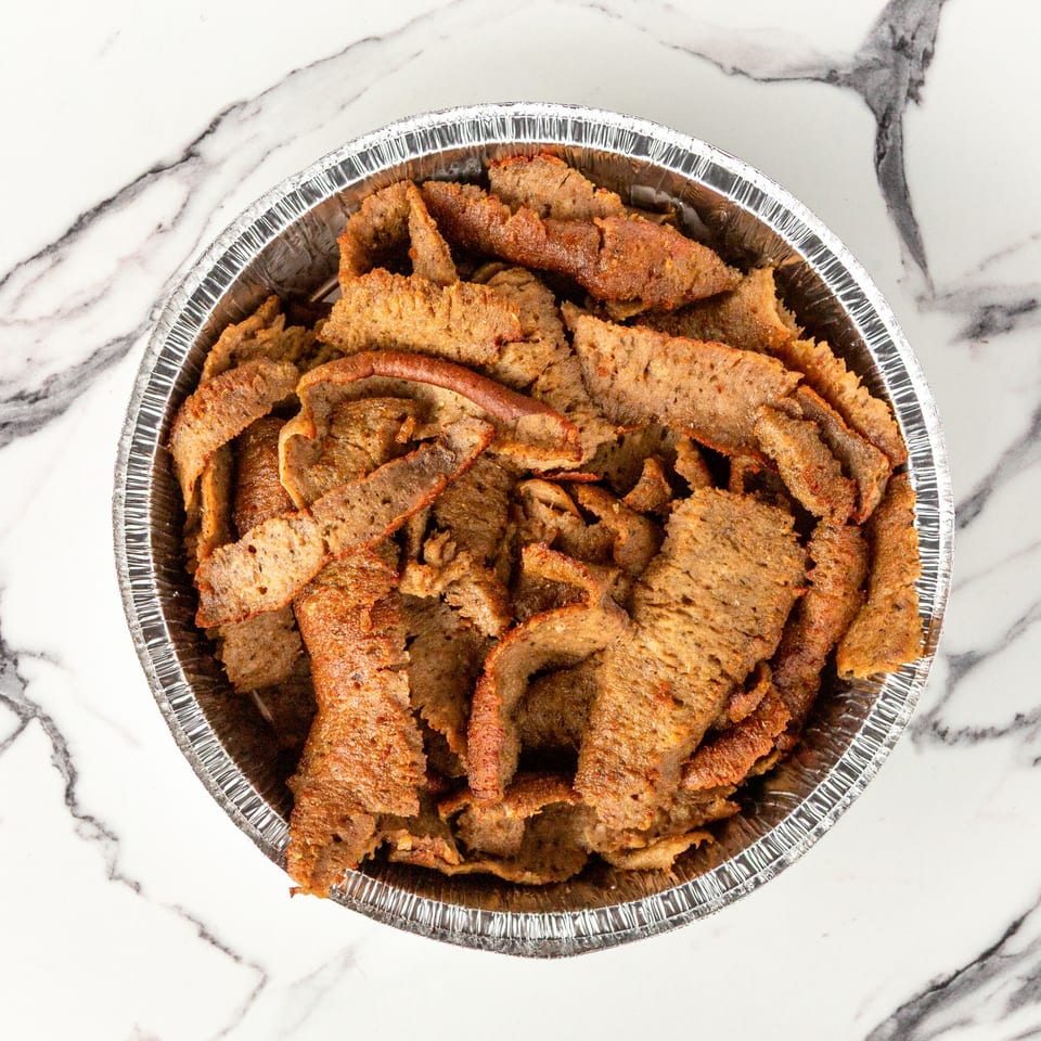 A foil container filled with thinly sliced, seasoned gyro meat on a white marble surface.