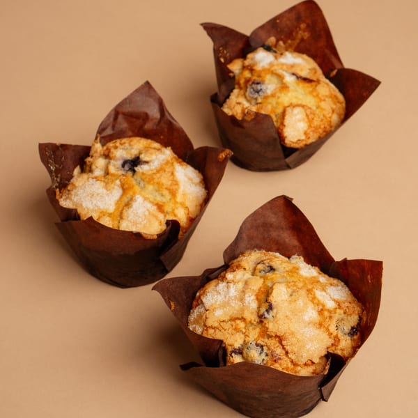 Three blueberry muffins with brown paper liners and a sugar topping on a tan surface.